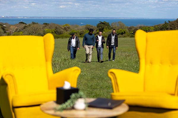 Two yellow chairs and a coffee table representing a therapist's office are set up on a Dorset hillside, four men are walking towards them across a field. In the background are trees and a view of the sea.